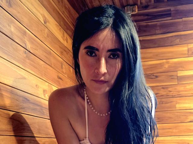 JainaRogers has long black hair, brown eyes, and a pearl necklace. She's wearing a white strap top, showing her small tits. She's leaning against wooden walls.