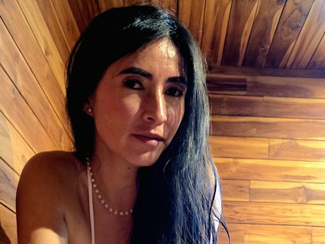 JainaRogers has long black hair, dark eyebrows, and wears a pearl necklace, with a serious expression, looking slightly to the side, in a wooden room.