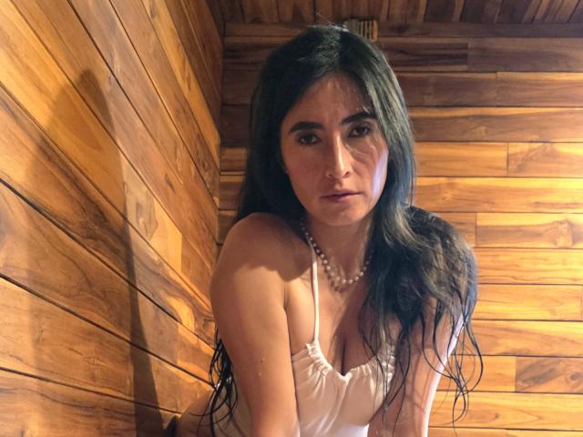 JainaRogers has wet long black hair, wearing a white dress with a pearl necklace, showing her tits, looking serious, in a wooden room.