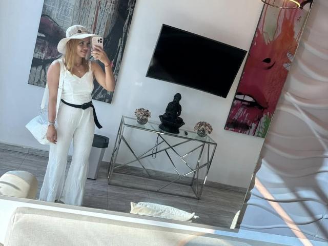 KataSofia with blonde hair, white hat, white sleeveless top, and white pants, holding phone, black belt, black handbag, standing in front of mirror, smiling, showing her ass.