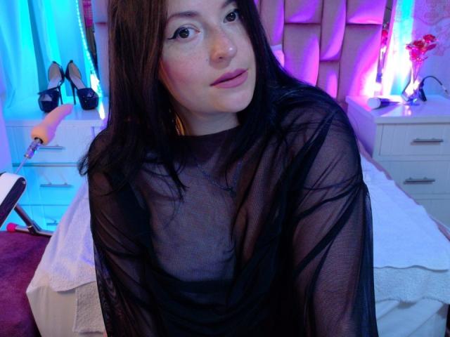 VaaleryStark with long black hair, wearing a sheer black top, showing her tits through the fabric, sitting in a bedroom with pink and blue neon lights.