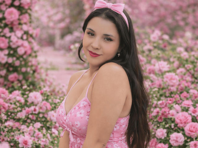 RosieTurner with pink bow headband, long black hair, pink floral dress, and pink lipstick, showing her tits in a pink flower-filled garden. She's smiling with red lipstick.