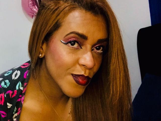 ShamanthaFire has brown hair, red lipstick, and pink eyeshadow with white winged eyeliner. She's wearing a black, kiss-patterned top, looking at the camera with a slight smile.