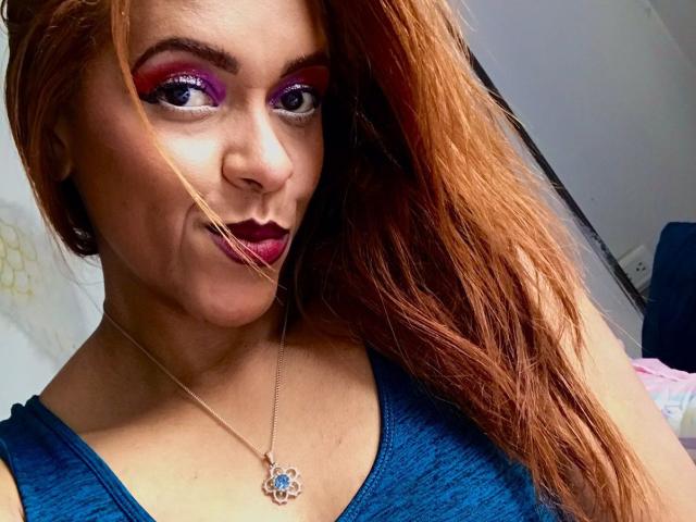 ShamanthaFire with red eyeshadow, purple lipstick, and blue top, showing off her tits with a nose hair. She's got a pendant necklace and long, messy red hair.