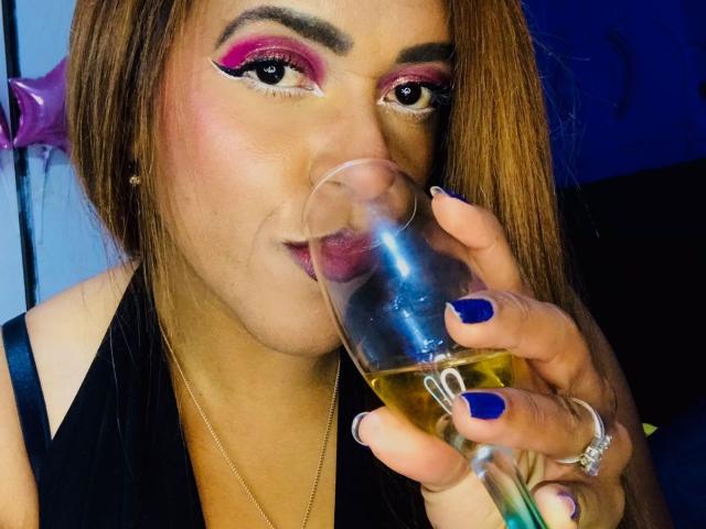 ShamanthaFire with pink eyeliner, blue nail polish, drinking from a glass of champagne, showing her ring, and wearing a black top, has long brown hair and a cute face.