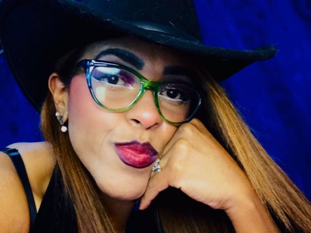 ShamanthaFire with green and blue glasses, pouty pink lips, brown hair, black hat, and black top, resting chin on hand, showing off a diamond ring.