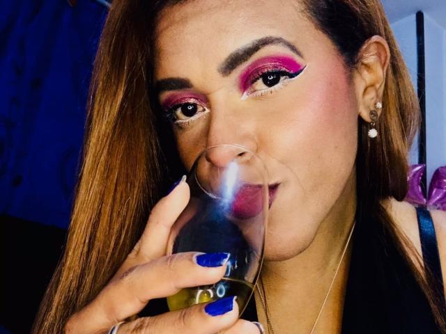ShamanthaFire with pink and purple eyeshadow, drinking from a glass, blue nails, long brown hair, earrings, and a black top, slightly smirking.