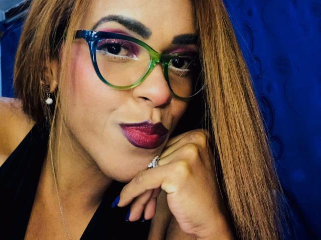 ShamanthaFire with green-blue glasses, pink-purple eyeshadow, red lipstick, brown hair, black top, diamond ring, hand on chin, blue background, looking at camera, smiling slightly.