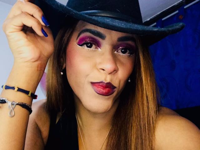 ShamanthaFire with blue nails, wearing a black hat, purple eyeshadow, red lipstick, and a black top, pouting her lips. She's holding her hat with her right hand.