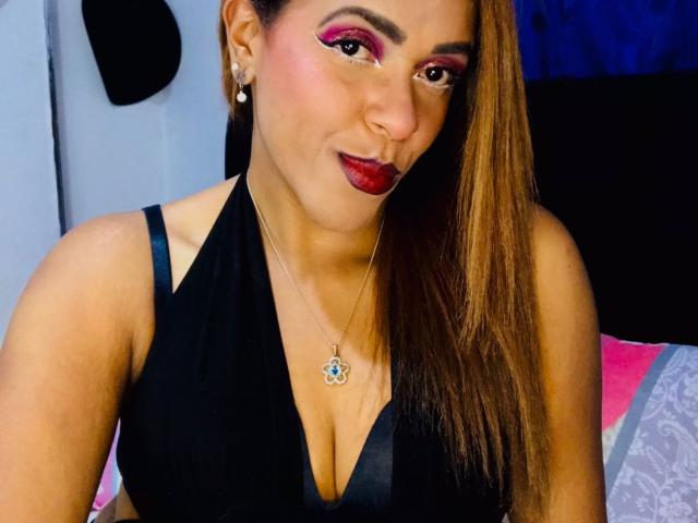 ShamanthaFire with long brown hair, pink eyeshadow, red lipstick, black halter top, showing her tits, necklace with blue flower, smiling, in a bedroom.