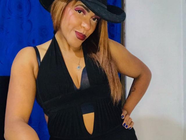 ShamanthaFire with tan skin, long brown hair, black hat, pink eye makeup, red lipstick, black halter dress, showing cleavage, hands on hips, blue nails, standing confidently.