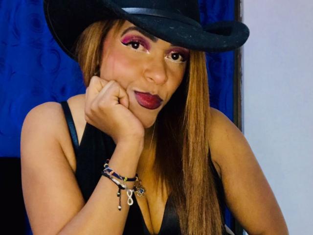 ShamanthaFire with long brown hair, wearing a black cowboy hat, black sleeveless top, and red makeup, resting her chin on her hand, smiling with red lipstick.
