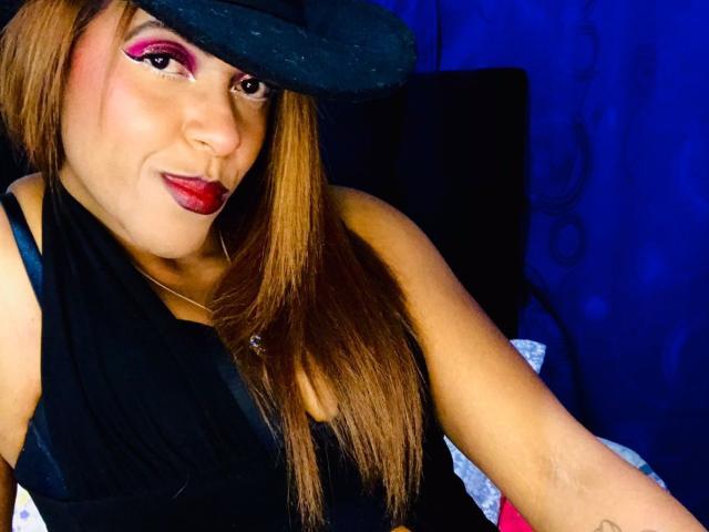 ShamanthaFire with long brown hair, black sleeveless top, red lipstick, pink eyeshadow, black hat, smiling, blue background, visible tattoo on right arm.