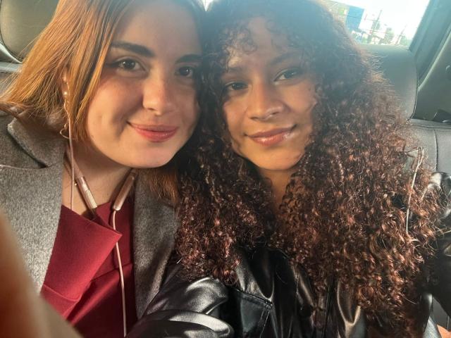 SofyViky with red hair and SofyViky with curly brown hair, both smiling, sitting in a car. SofyViky in a gray coat, red shirt, black leather jacket.