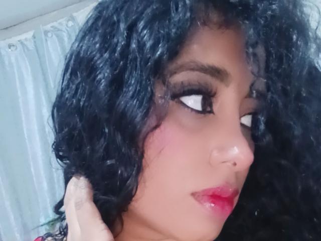 MariaCatalina's face with curly black hair, brown eyes, and red lipstick, looking to the side. She has a nose piercing and a tattoo on her hand.