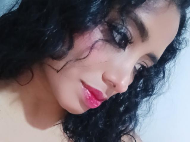 MariaCatalina with curly black hair, pink glossy lips, and dark eyeliner, looking to the side with a slight smile, showing her cute nose and soft skin.