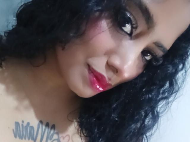 MariaCatalina with curly black hair, glossy pink lips, and dark eyes, showing a slight smile, has a tattoo on her chest, and is looking up at the camera.
