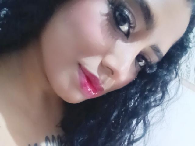 MariaCatalina's face, showing her dark curly hair, pink glossy lips, and large brown eyes with thick eyelashes, looking slightly to the side with a subtle smile.