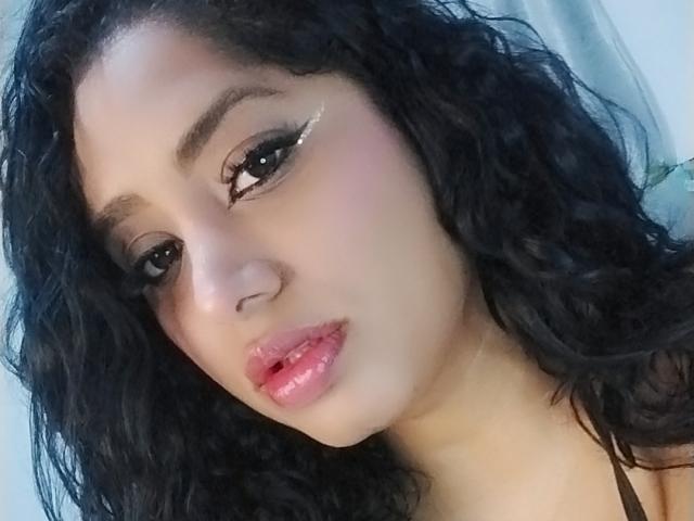 MariaCatalina has curly black hair, brown eyes with winged eyeliner, and shiny pink lips. Her skin is light brown, and she's looking slightly to the left.