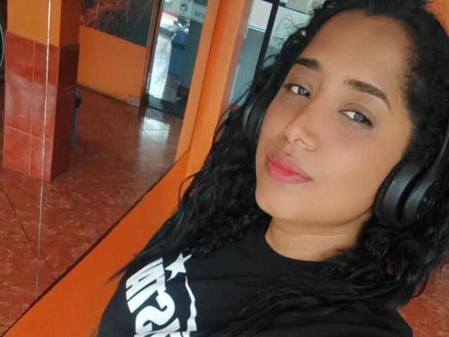 MariaCatalina with curly black hair, wearing black headphones, black shirt, and pink lipstick, smiling slightly at the camera, showing her nose and lips. Her brown eyes are looking forward.