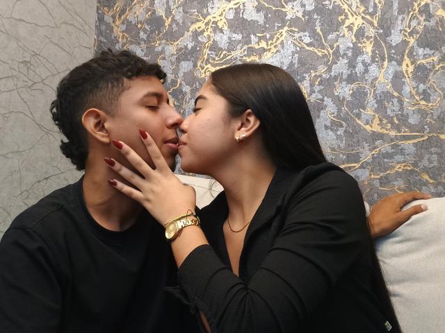 SofiAndLuis are kissing, her hand with red nails on Luis's cheek, both in black clothes. She's wearing a gold watch and earrings. Luis has curly hair.