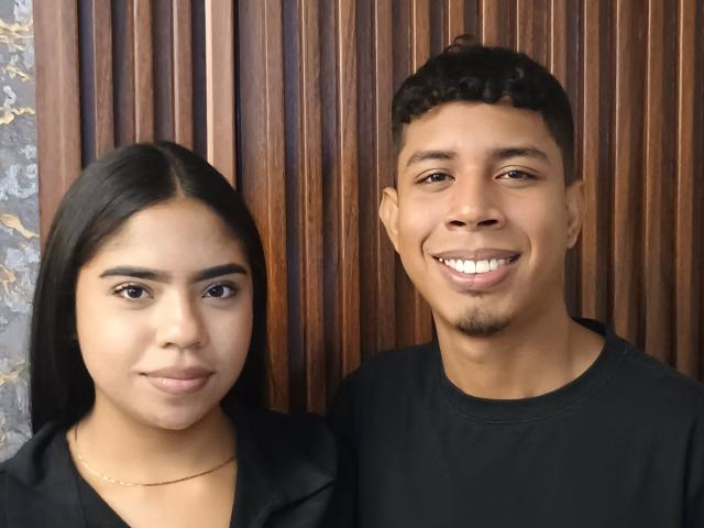 SofiAndLuis has tan skin, dark hair, and is smiling with white teeth. Luis has tan skin, short curly black hair, and is also smiling. Both wear black shirts.