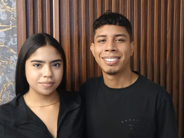 SofiAndLuis (her) with long black hair, wearing a black shirt, and Luis (him) with short black hair, smiling, wearing a black shirt, standing together.