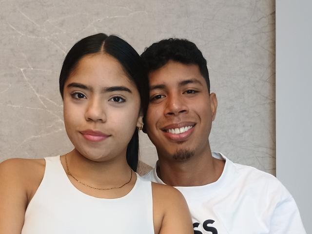 SofiAndLuis, a cute Latina chick with long black hair and small tits, wearing a white tank top, stands next to a smiling, dark-skinned Latino dude in a white tee.