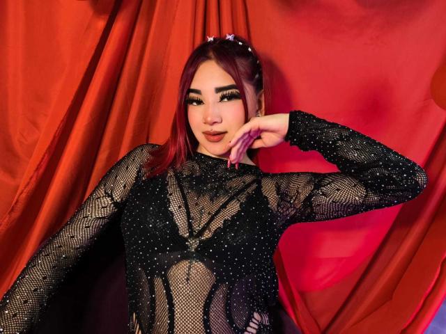 MiilaRouge with red hair, wearing a black, sheer, sparkly top, showing her small tits through the mesh, hand on cheek, red curtain background, looking seductive.