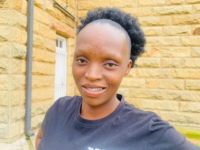 YoguetaLips with a bald top, curly black hair, brown skin, smiling, wearing a black shirt, standing in front of a yellow stone wall.