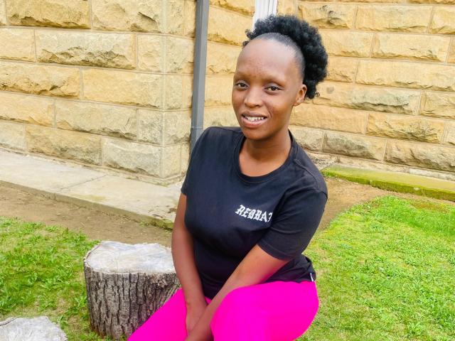 YoguetaLips with dark skin, small round tits in a black t-shirt, bright pink pants, sitting on a tree stump, smiling, short curly black hair, brick wall in background.