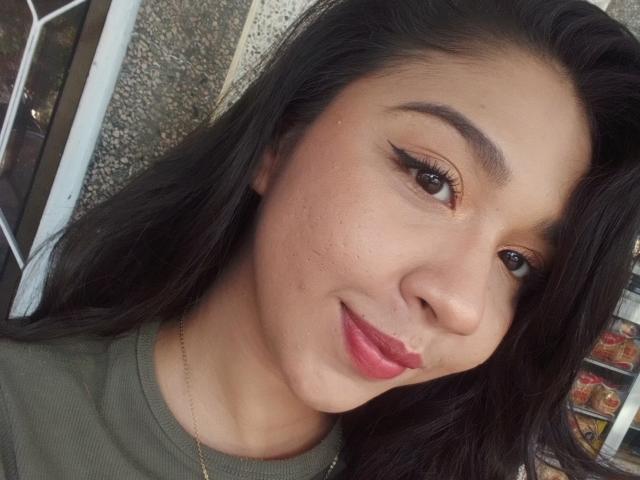 NanaMilan's face, dark hair, brown eyes, winged eyeliner, pink lipstick, green top, subtle freckles, slight smile, gold necklace, medium skin tone, casual selfie.