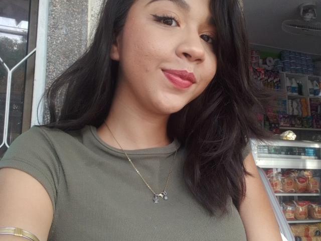 NanaMilan with medium brown skin, black wavy hair, green shirt, gold necklace, pink lipstick, and subtle eyeliner, smiling softly, showing slight pimple on her cheek.