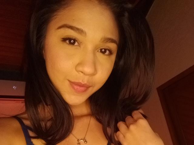 NanaMilan has medium brown skin, black hair, and brown eyes with a slight pout, wearing a blue top and a gold necklace, holding her hair.