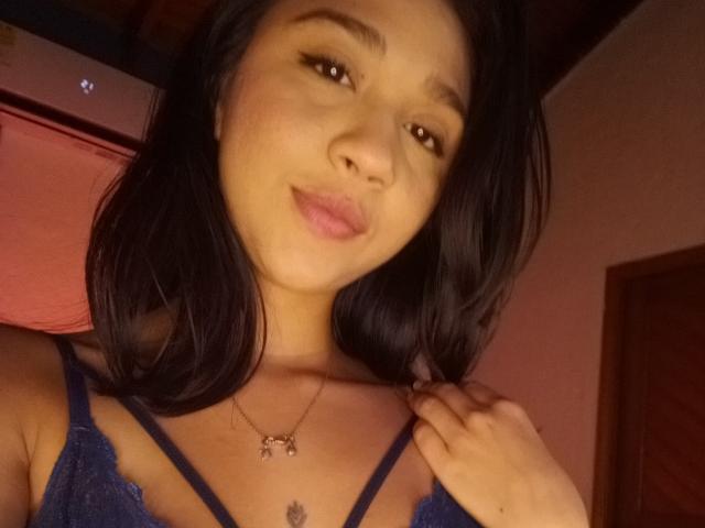 NanaMilan with dark hair, brown eyes, and a slight smile, wearing a blue lace top, holds her hair, showing a necklace with a small charm and a tattoo.