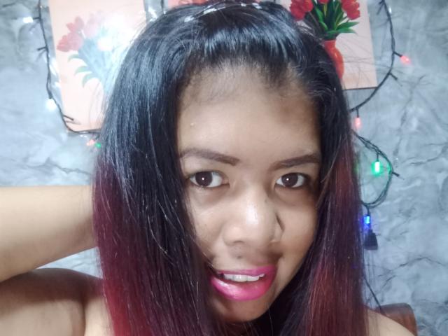 MariahSquirting69 has dark brown skin, black hair with red tips, and is smiling with pink lipstick, looking at the camera, with a Christmas light string in the background.