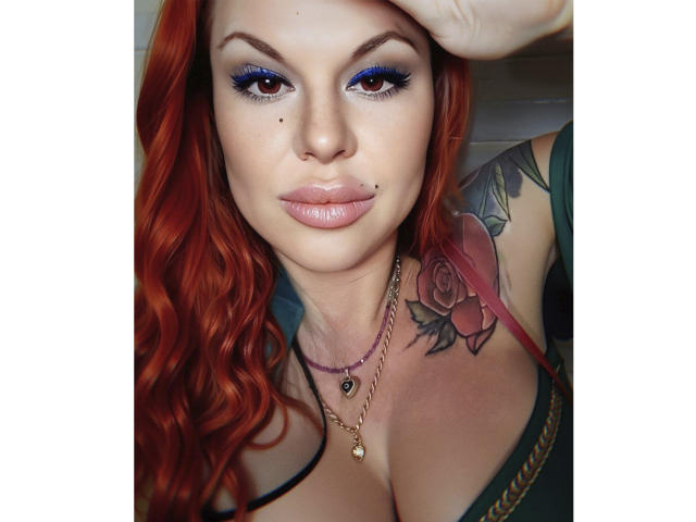 MissTwinkleX with red hair, blue eyeshadow, and a rose tattoo on her chest, wearing a green top, showing her large, perky tits, and a nose piercing.