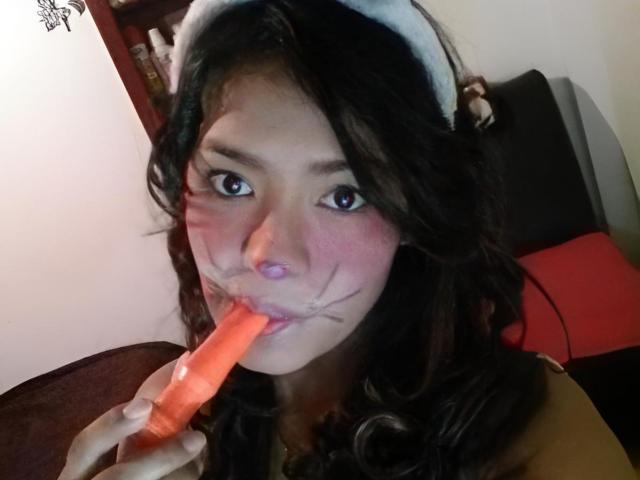 ArianithaXox, con i capelli scuri e ondulati, indossa una fascia bianca e sta succhiando un dildo rosa con delle macchie bianche sul viso. I suoi occhi sono spalancati e sta guardando la fotocamera..