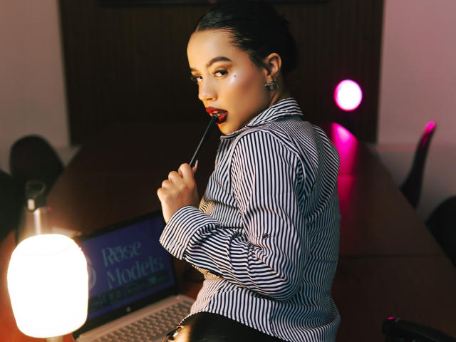 WendyJames with dark skin, red lipstick, and striped shirt, biting a red-tipped pen, looking over shoulder, laptop on desk, pink and white lights, shiny earrings.