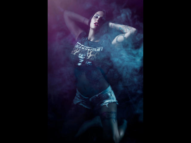 XANIA-WET with tattoos, wearing a crop top and denim shorts, arms raised, smoke around, dark purple and blue lighting, red lipstick, and a confident, sexy pose.