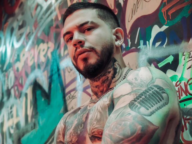 AlessioMoonOfficial, a heavily tattooed man with a short beard and ear gauges, stands against a graffiti-covered wall. His tattoos are intricate, featuring a large skull on his right shoulder and a detailed chest piece. He has a serious expression, looking slightly to the left. His dark hair is styled in a buzz cut. The wall behind him is filled with colorful graffiti, including green, red, and blue spray paint. The image has a gritty, urban feel with sharp contrasts between AlessioMoonOfficial's tattoos and the colorful graffiti. The lighting is dynamic, with red and green hues.