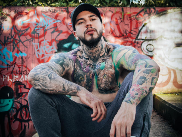 AlessioMoonOfficial, shirtless, crouches in front of a graffiti-covered wall. He's heavily tattooed with colorful designs on his chest, arms, and neck. He wears a black cap and gray pants. His beard is short and dark. His tattoos include a green-eyed face on his chest and a mix of geometric and illustrative designs on his arms. The graffiti in the background is chaotic with red, black, and blue tags. Natural light highlights his tattoos and facial features. His expression is relaxed, looking slightly upward. The overall composition emphasizes his tattoos and street-style appearance.