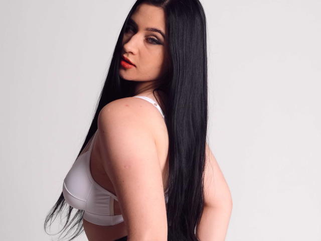 MaryLori, a white woman with long, straight black hair, stares over her shoulder. She wears a white bra with thin straps, revealing her fair skin. Her red lipstick contrasts sharply with her pale complexion. The image focuses on her upper body and face, highlighting her sharp, dark eyebrows and subtle, smoky eye makeup. Her hair is sleek, reaching down her back. The photograph is simple, with a plain white background, emphasizing her striking features and bold red lips.