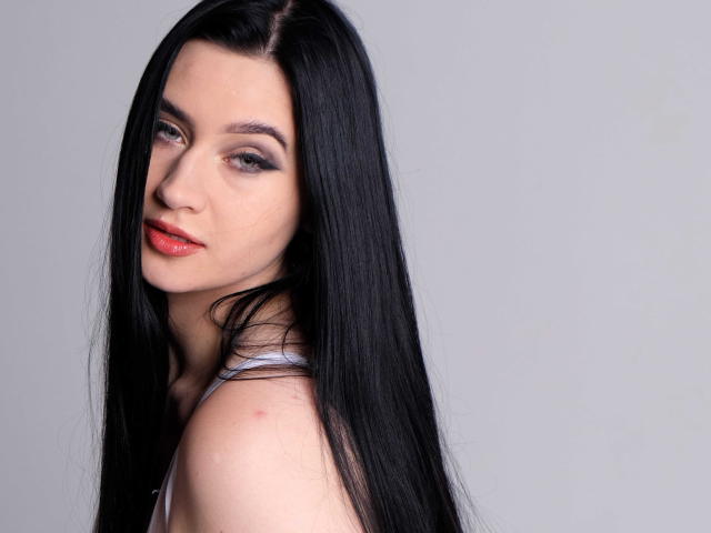 MaryLori, a fair-skinned woman with long, straight black hair, is shown in a close-up portrait. She has subtle, dark eye makeup and red lipstick. Her right shoulder is visible, showing a small red mark. She looks over her right shoulder with a slight, confident smile. The image is sharply focused, highlighting the smooth texture of her hair and skin. The background is a plain, light grey, keeping all attention on MaryLori. She has a slender build and a composed, slightly sultry expression.