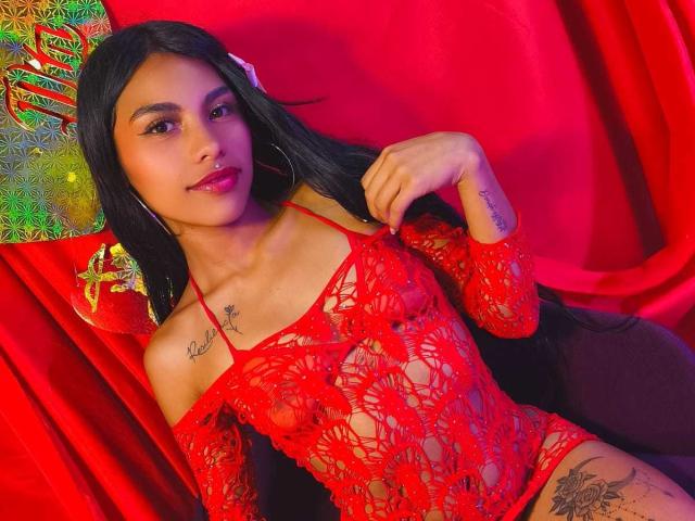 ClarisCampbell with long black hair, red lace bodysuit, hoop earrings, nose ring, tattoos, and red lipstick, sitting on a red chair, showing small tits and a tattooed thigh.