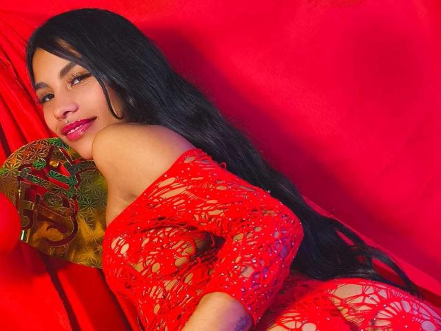 ClarisCampbell with long black hair, red lace dress, and pink lipstick, lying on red fabric, smiling, holding colorful pillow, red background, looking over shoulder, showing ass.