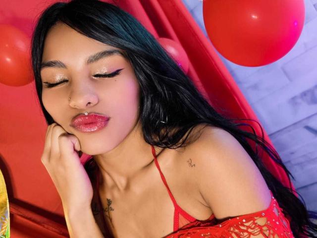 ClarisCampbell with long black hair, red lipstick, glittery eyeliner, red off-shoulder top, and red balloons in the background, touching her cheek with her hand, eyes closed.