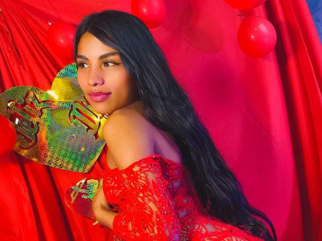 ClarisCampbell with long black hair, wearing a red lace dress, holding a colorful, ornate mask, and red balloons in the background. Her pink lipstick matches the vibrant red.