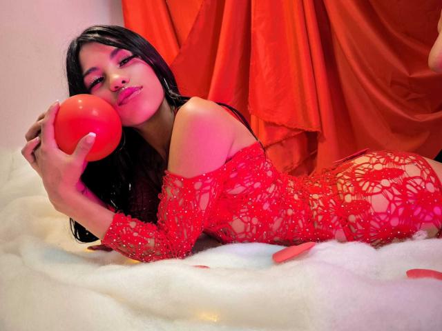 ClarisCampbell with long black hair, red lace dress, and red lipstick, holding a red ball, lying on white foam, red drapes in background, pink eye makeup.