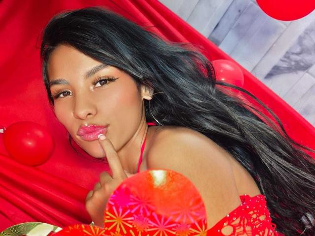 ClarisCampbell with long black hair, red lips, and a finger in her mouth, wearing red lace, surrounded by red balloons, and holding a red fan with pink patterns.